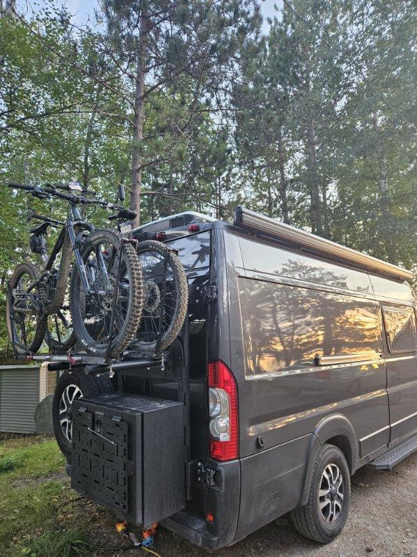 parked in a pine forest at sunset. Its lights glow.|Explore with an RV rental! Dark grey camper van parked on a scenic gravel road amidst lush trees and blue sky.|Black camper van on dirt road