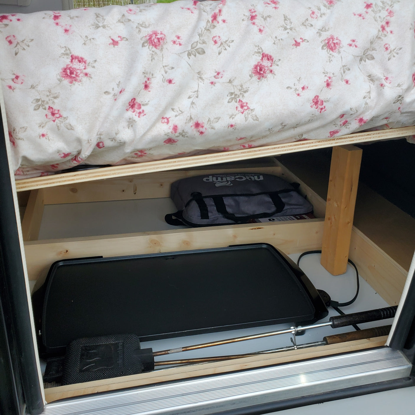 Compact RV rental storage: floral bed above, nuCamp bag, electric griddle, and roasting stick below. Efficient space.