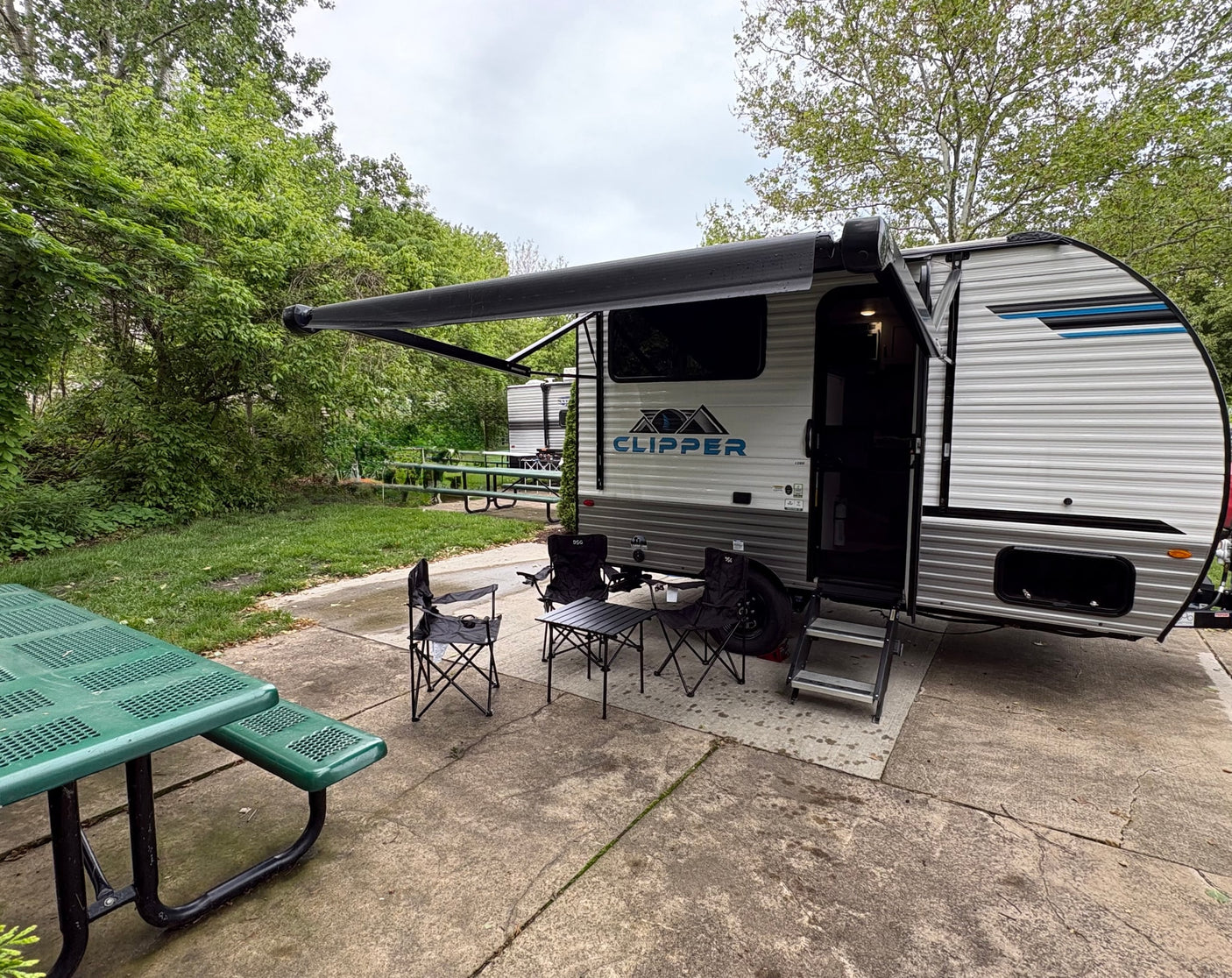 A modern RV rental travel trailer is set up at a peaceful campsite with an extended awning, chairs, and a picnic table.