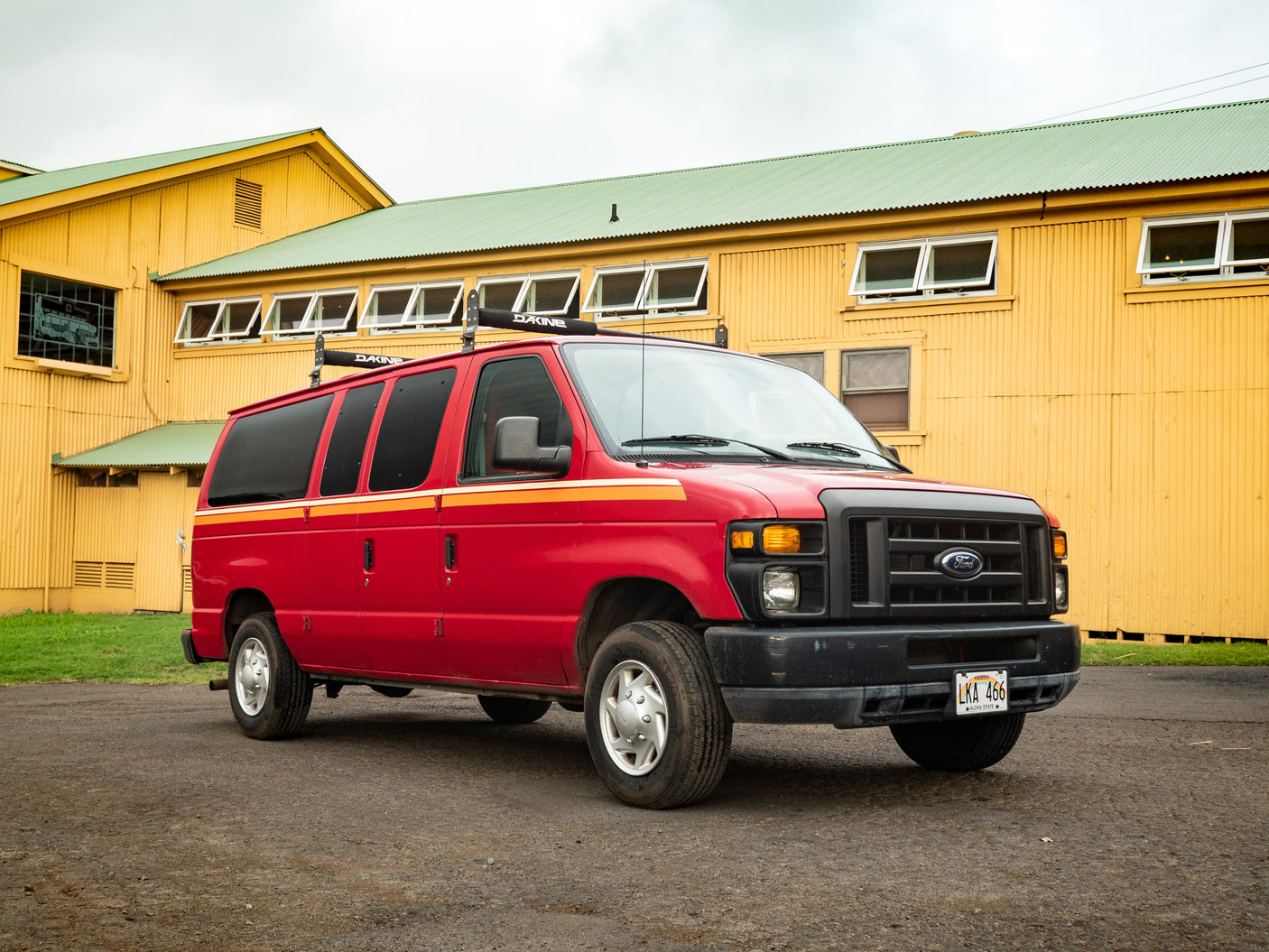 A vibrant red Ford E-Series camper van, an RV rental with Dakine racks, parked by a sunny yellow Hawaiian building.