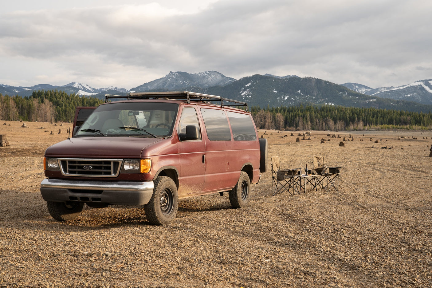 Explore nature! Maroon RV rental van with camping chairs on a dry, stump-filled landscape, mountains beyond.