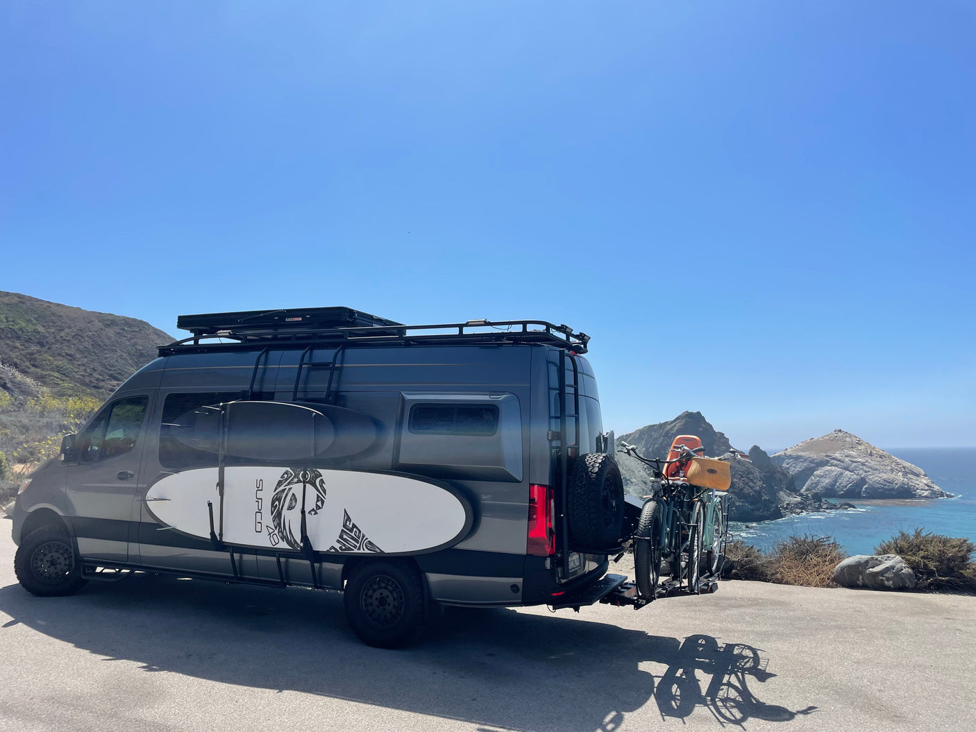 Custom RV rental van with surfboards and bikes by the scenic ocean and mountains. Ideal for adventure travel.