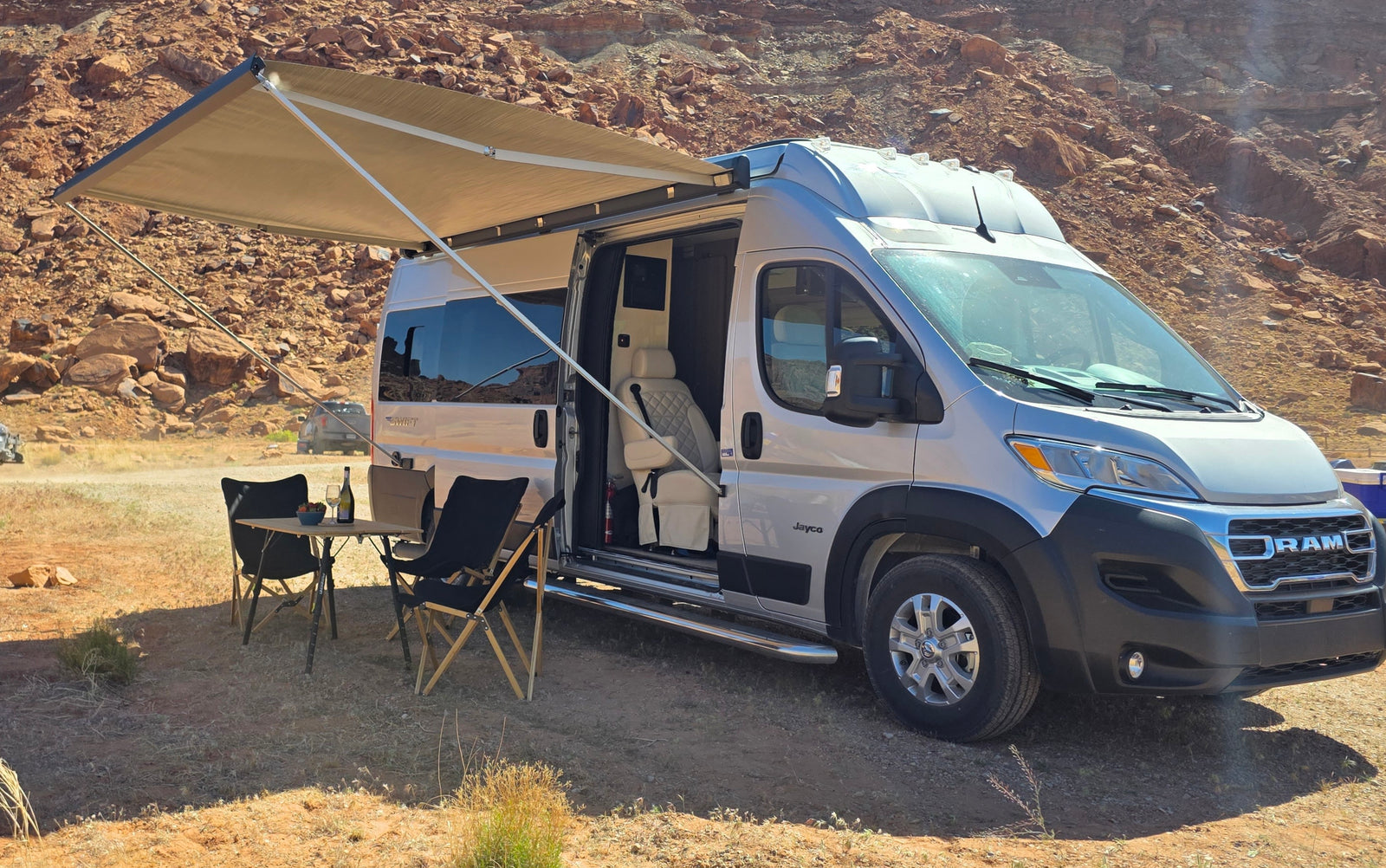 Desert boondocking with this RV rental. Camper van with open awning, chairs, and table set up in a rocky landscape.