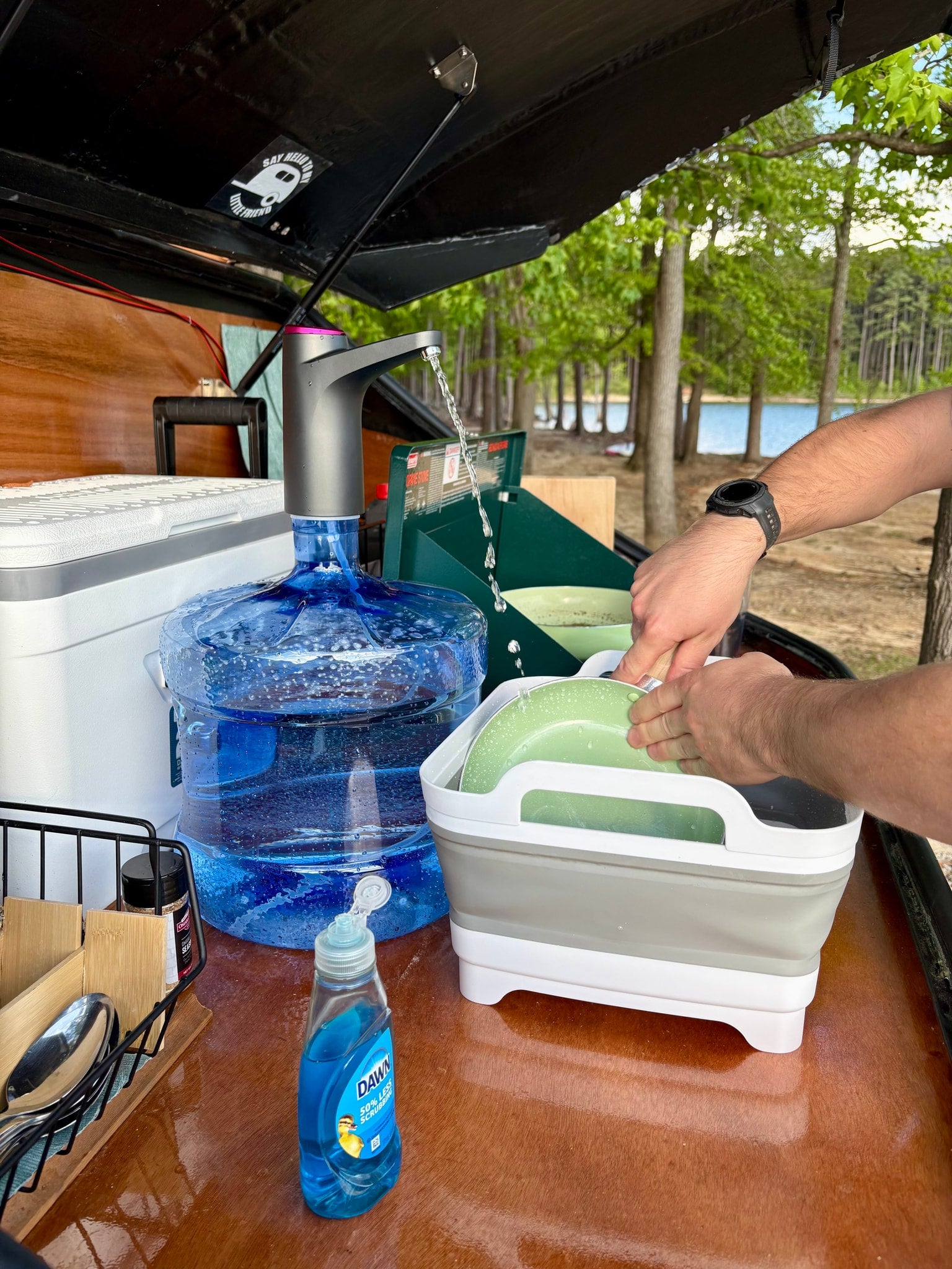 and power controls showing 13.9V.|An RV rental teardrop trailer's open hatch reveals an outdoor kitchen by a lake