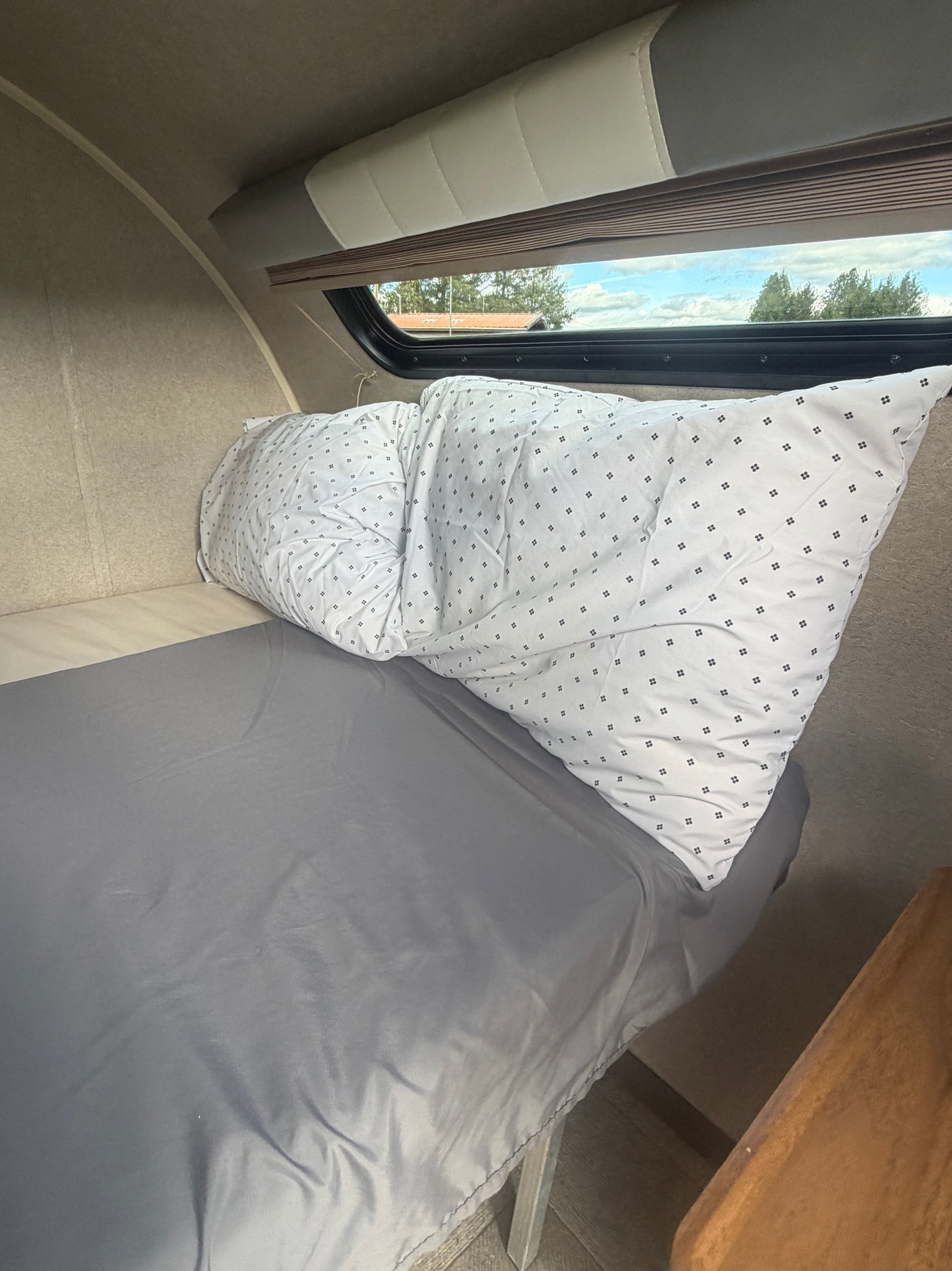 Inside an RV rental: A comfortable bed with grey sheets and a white patterned comforter, nestled under a window.