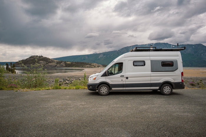 White camper van (rv rental) parked on gravel, facing a scenic lake, mountains & cloudy sky. Ready for adventure!