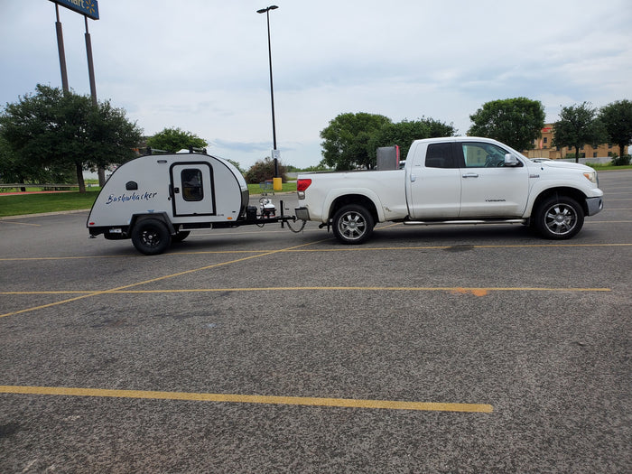 White pickup truck tows a compact Bushwhacker travel trailer, parked at a Walmart, an ideal RV rental.
