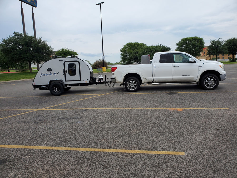 White pickup truck tows a compact Bushwhacker travel trailer, parked at a Walmart, an ideal RV rental.