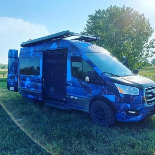 A blue camo-patterned camper van with open doors in a sunny field. Perfect for your next RV rental adventure!