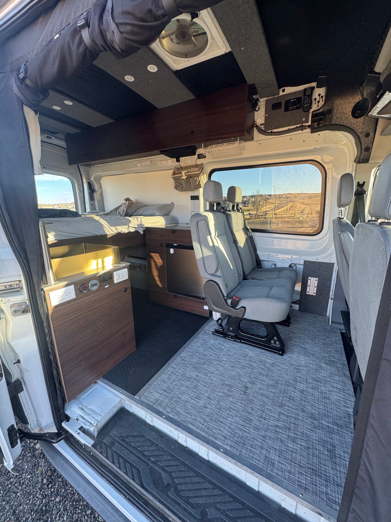 Inside this RV rental camper van, a bed, kitchen, and passenger seats are visible from the open side door.