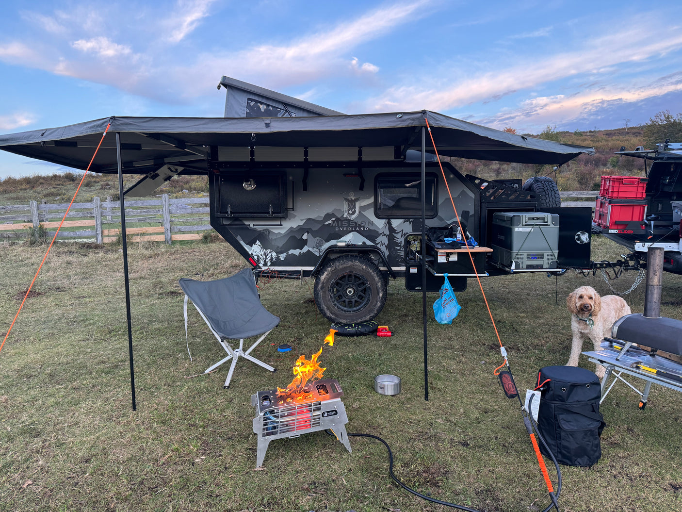 Scenic RV rental setup: overlanding trailer, cozy campfire, happy dog, and outdoor cooking station under an awning.