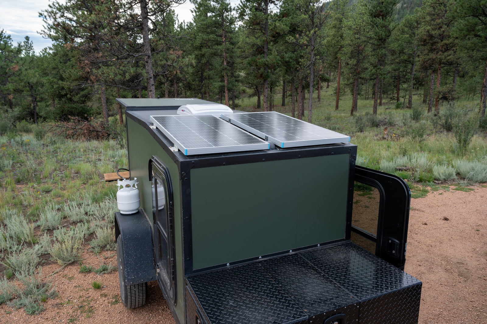 A dark green off-grid camper with roof solar panels and propane tank for RV rental camping in a pine forest.