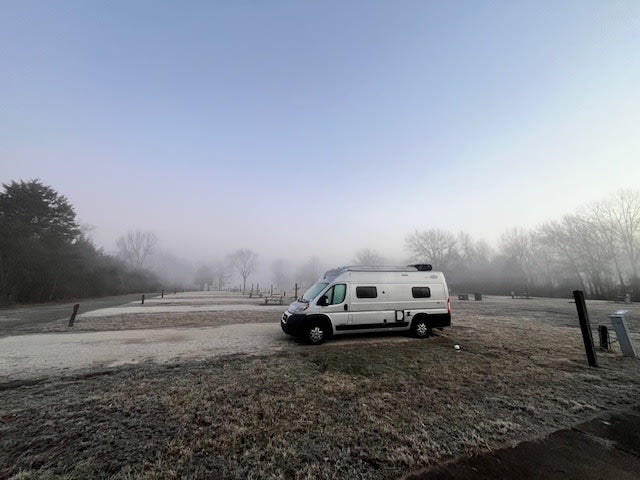 frosty morning at a quiet RV park. White camper van (RV rental) parked on frosted grass.|Coastal RV rental parked on sandy ground by palm trees