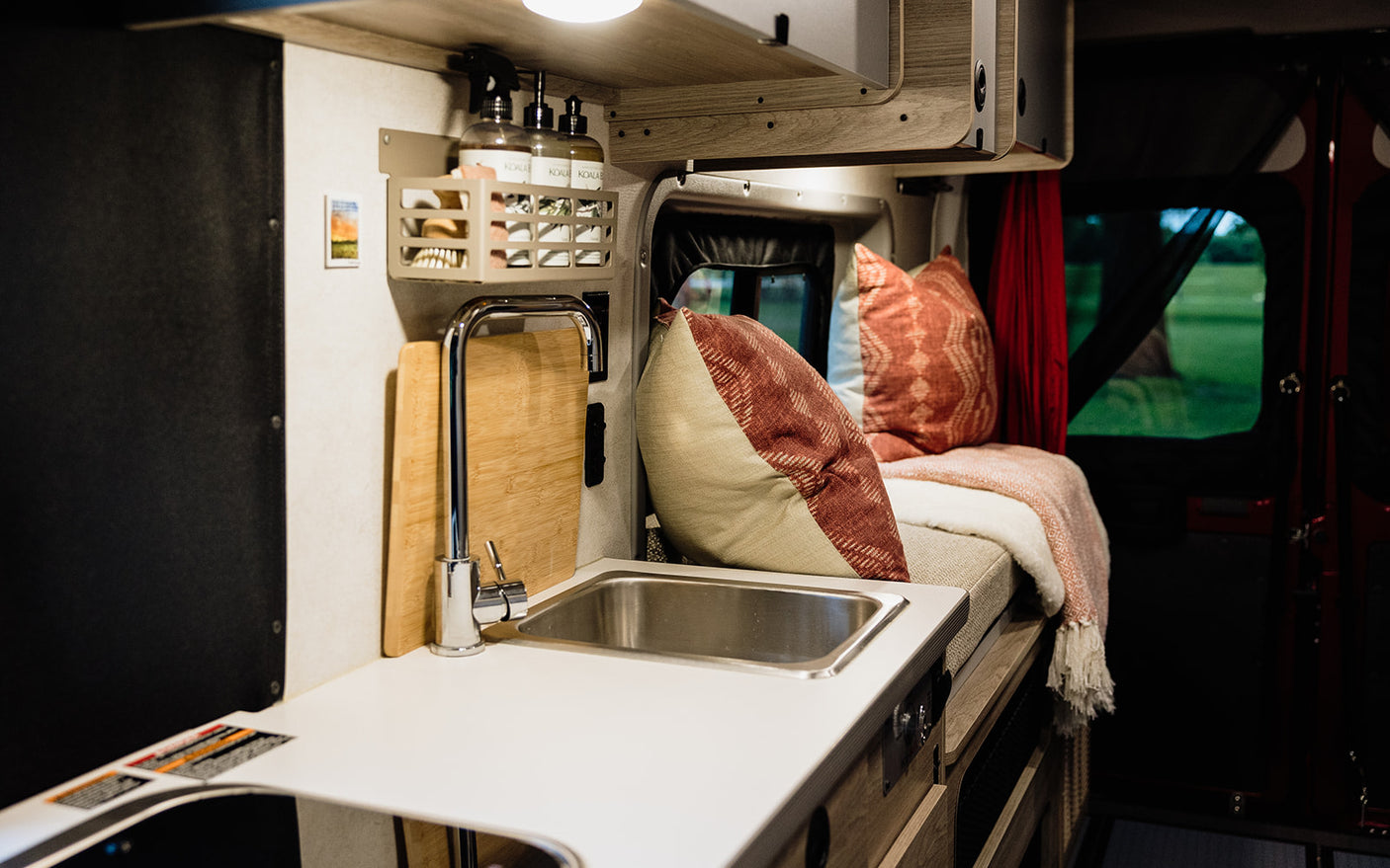 Inside a cozy RV rental: compact kitchen with sink, comfy seating, and a window revealing a green view.