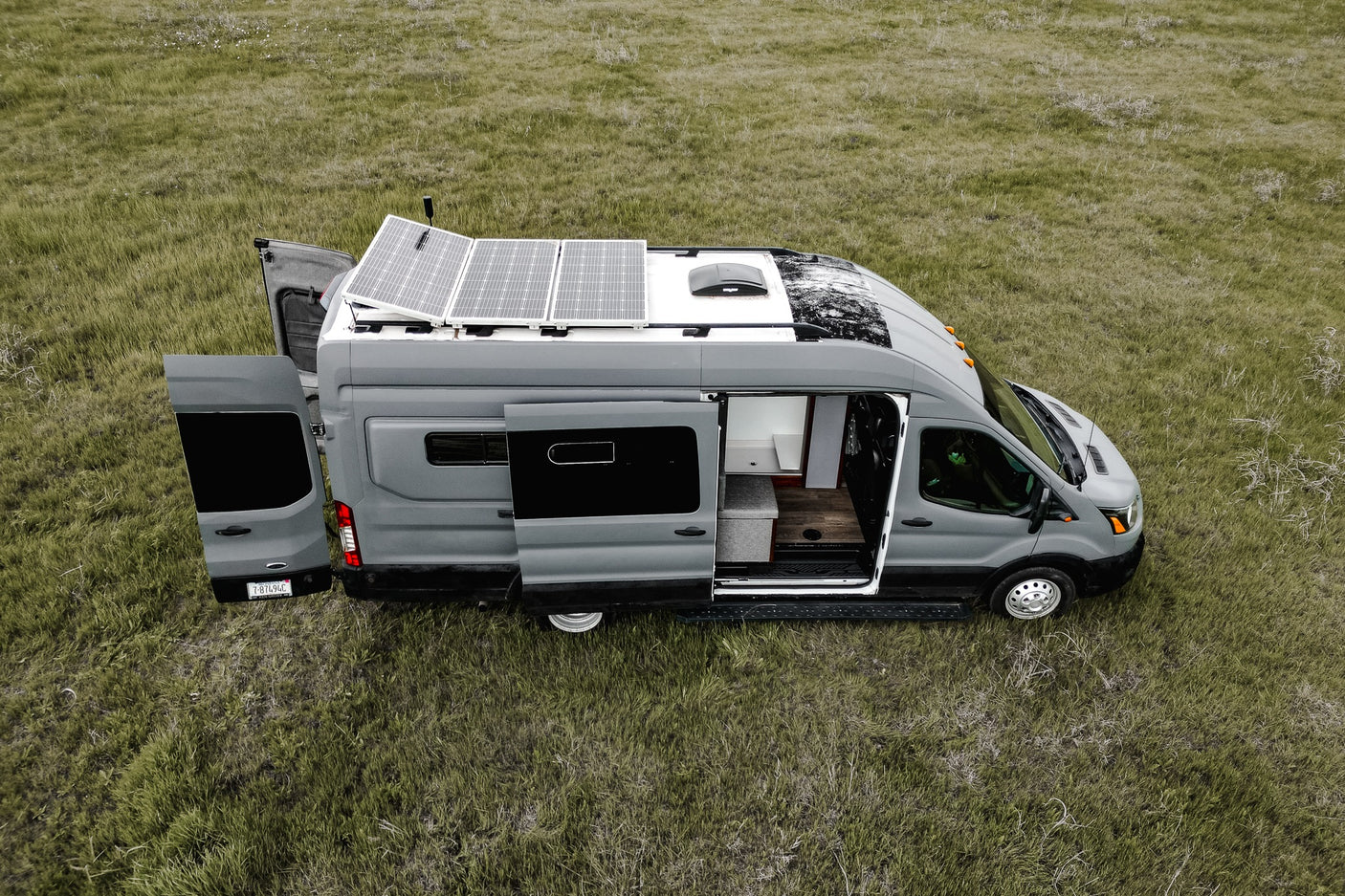 Explore the outdoors with this grey camper van RV rental, featuring solar panels and open doors revealing its cozy interior in a grassy field.