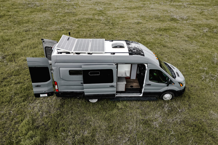 Explore the outdoors with this grey camper van RV rental, featuring solar panels and open doors revealing its cozy interior in a grassy field.