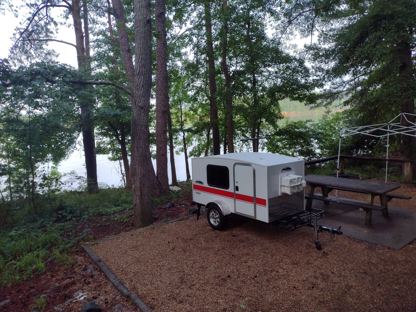 Small white & red teardrop RV rental at a serene lakeside campsite with trees & picnic table, perfect for outdoor adventures.