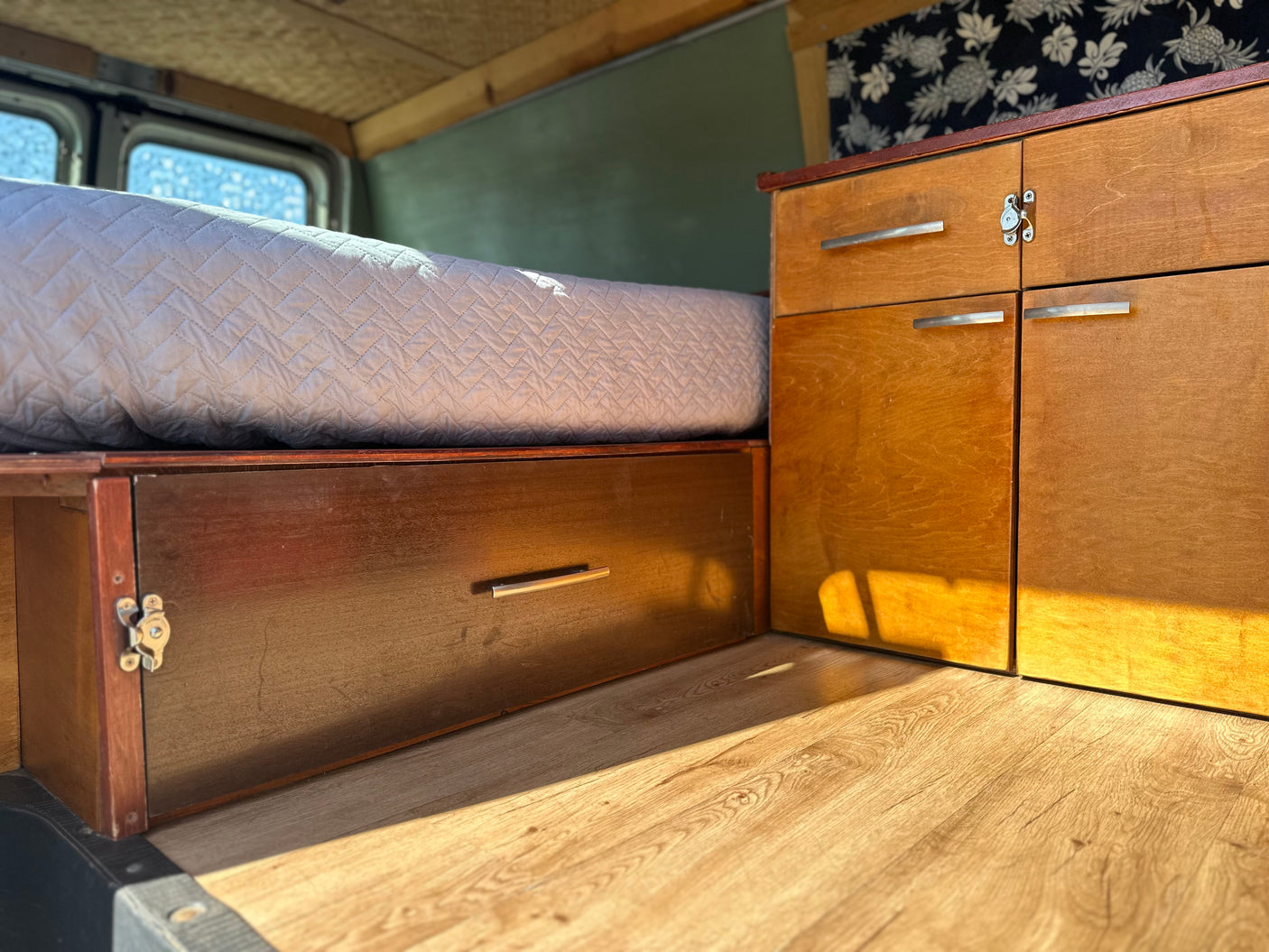 Bright RV rental interior: cozy bed with storage below, wood cabinets, and tropical wallpaper, bathed in sunlight.