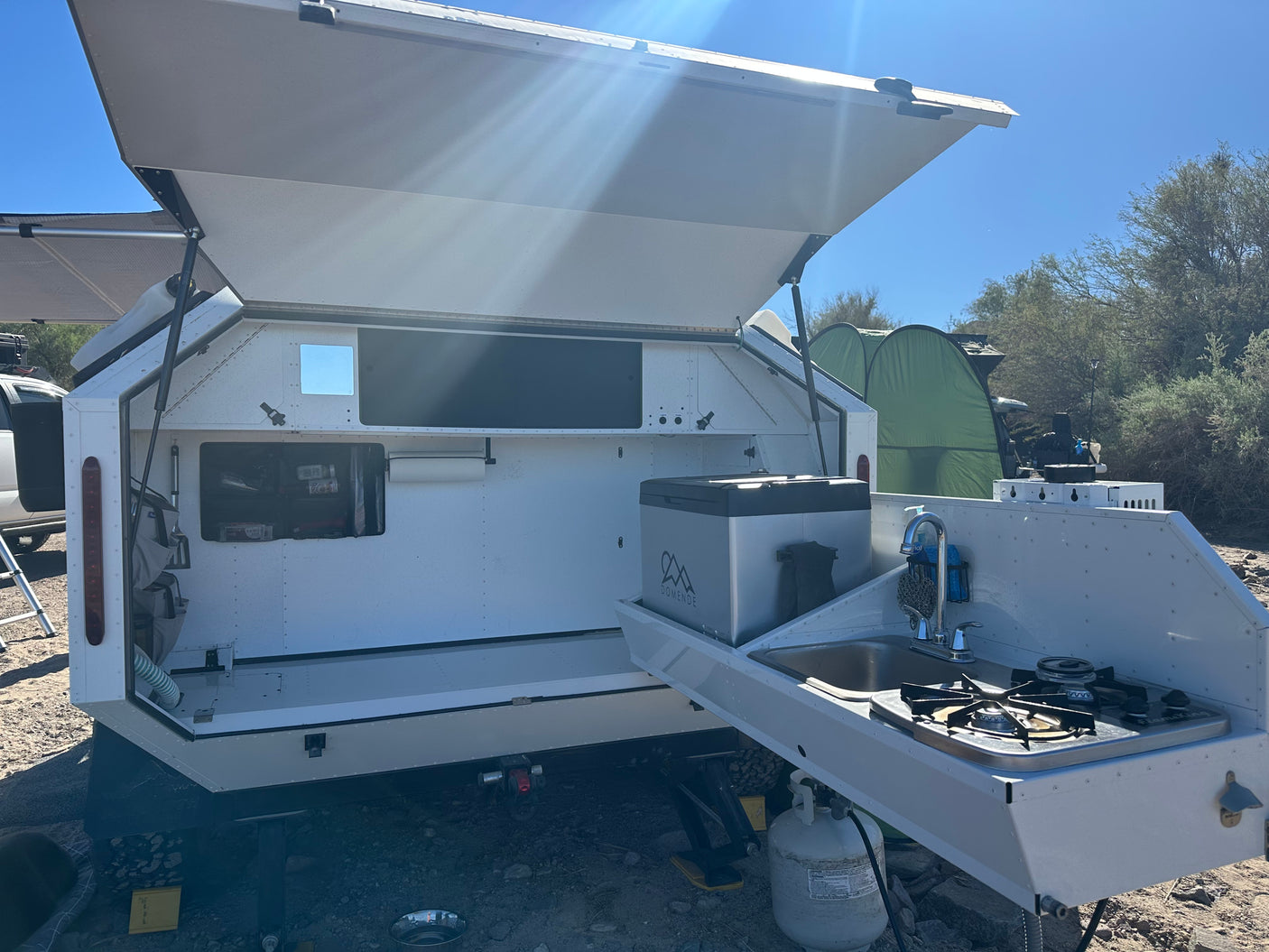 An RV rental features an open rear hatch revealing an outdoor kitchen with a sink, stove, and fridge for camping.