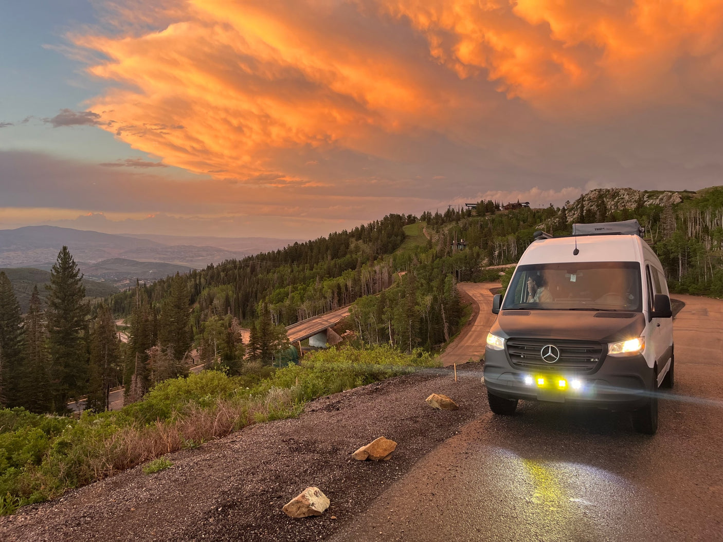 RV rental van on a mountain road at sunset, with glowing orange clouds over forested peaks. Scenic drive, perfect for outdoor adventure.
