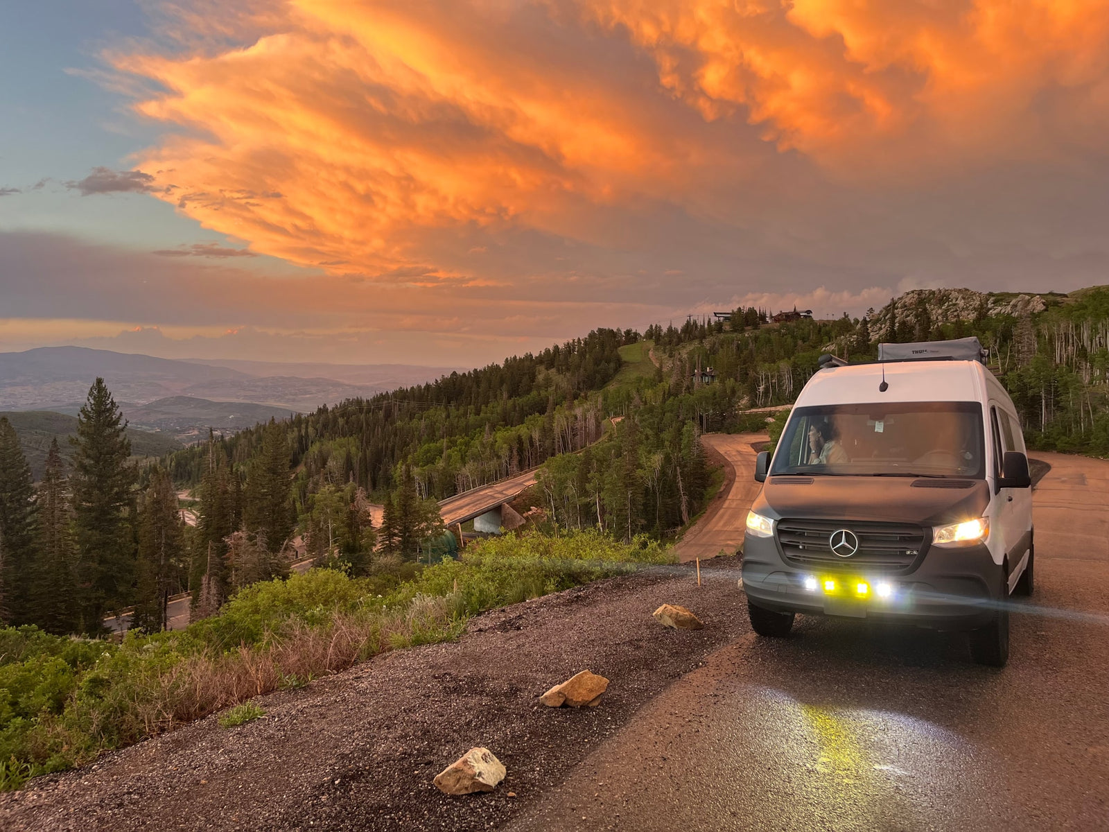 RV rental van on a mountain road at sunset, with glowing orange clouds over forested peaks. Scenic drive, perfect for outdoor adventure.