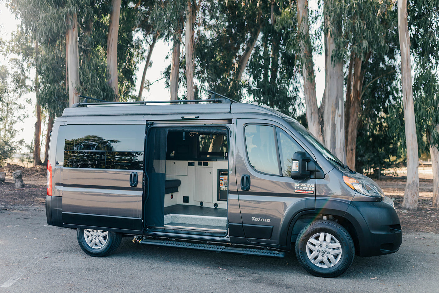 This gray Ram Promaster Tofino camper van RV rental shows its open side door and inviting interior setup, parked outdoors.