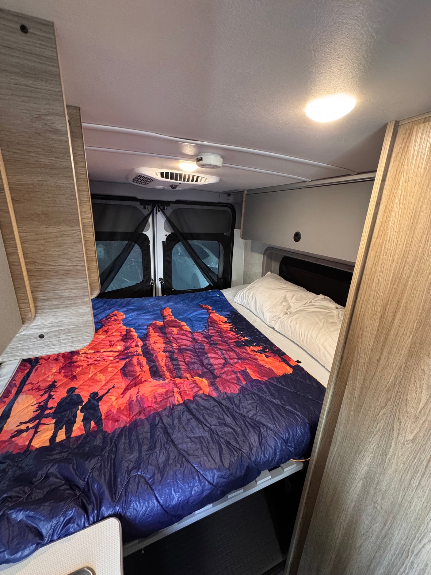 This cozy RV rental features a comfortable bedroom with a vibrant scenic blanket and overhead lighting for your travels.