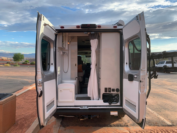Rear view of an RV rental camper van, doors open, showing an indoor shower, toilet, and power panel. Desert view.