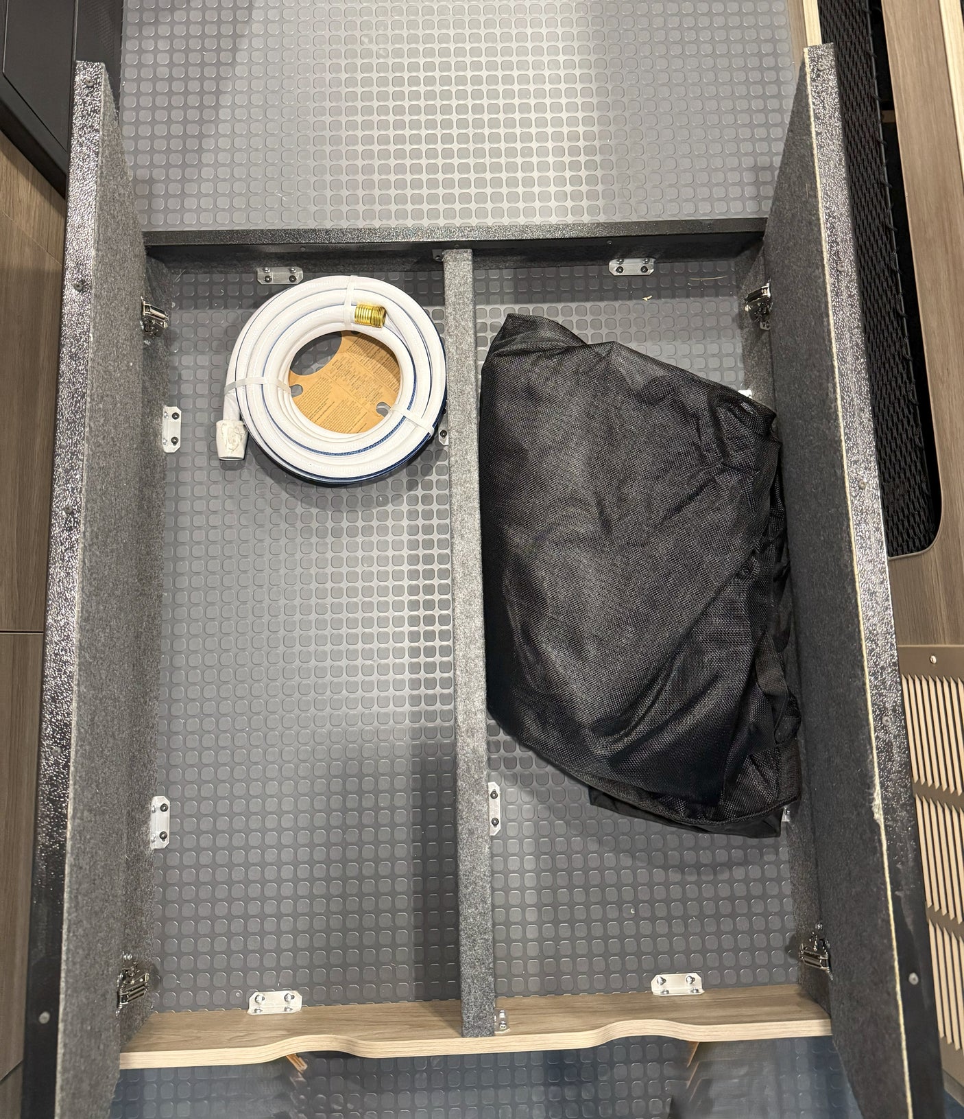 This rv rental's organized storage compartment holds a coiled white water hose and a black bag on a textured grey mat.