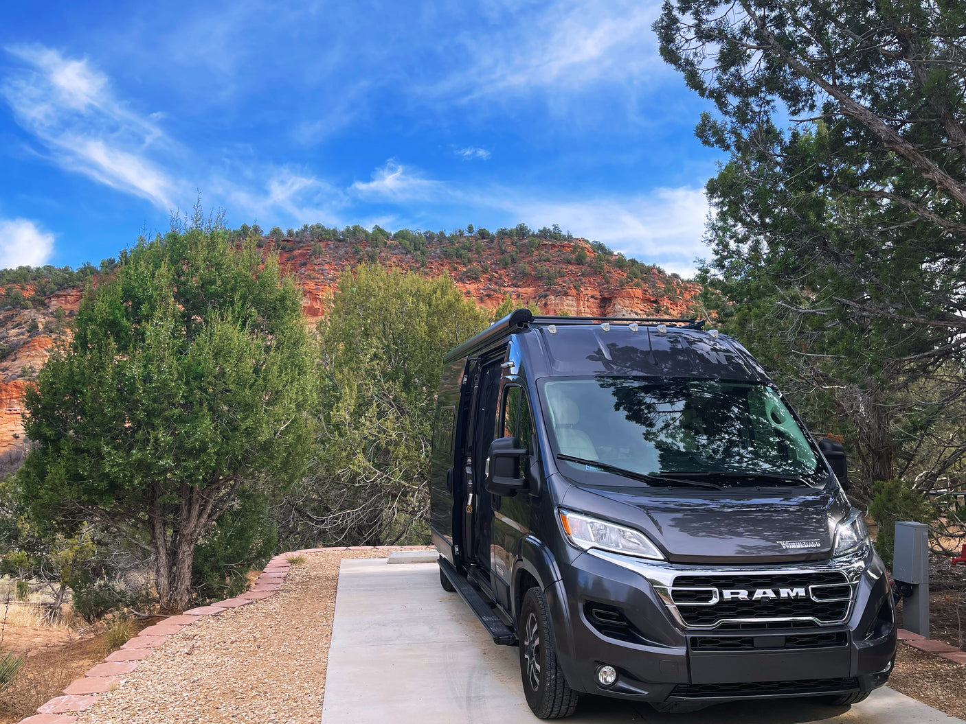 A modern grey rv rental camper van sits ready for adventure on a concrete pad, framed by stunning red rock formations and a vivid blue sky.