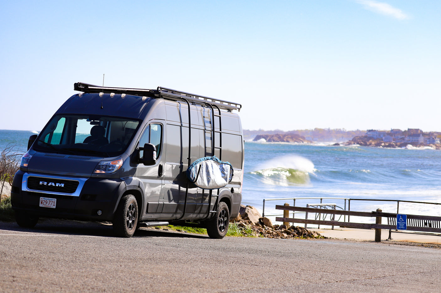 A dark gray camper van with a roof rack and surf bag overlooks crashing ocean waves. Ideal rv rental for beach adventures!