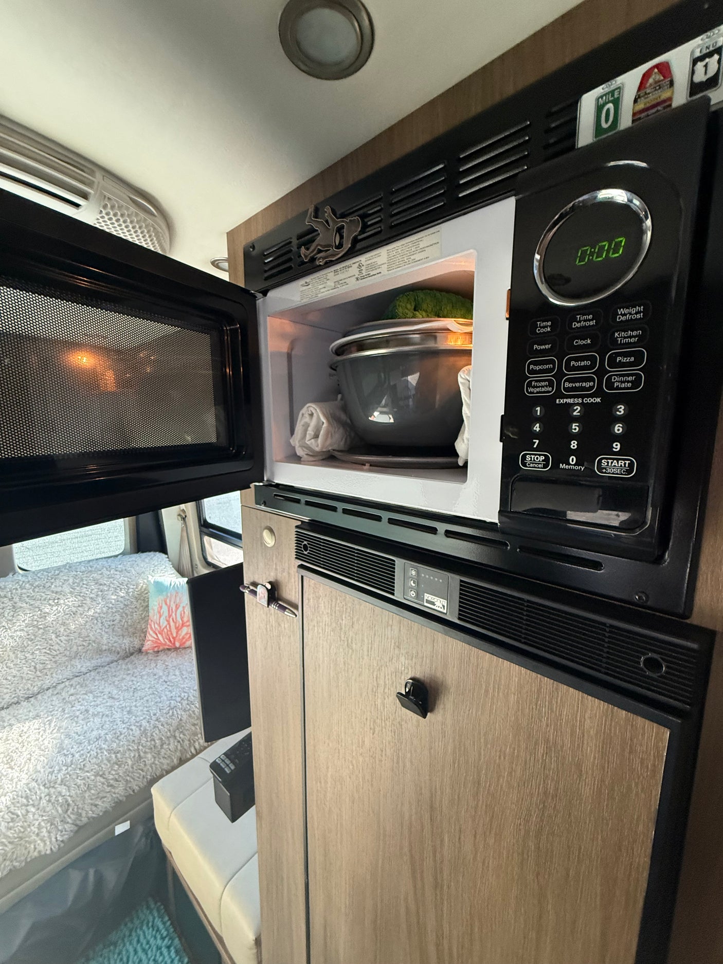 RV rental interior: A compact kitchen area featuring an open microwave with food, above a fridge, beside a cozy bed.