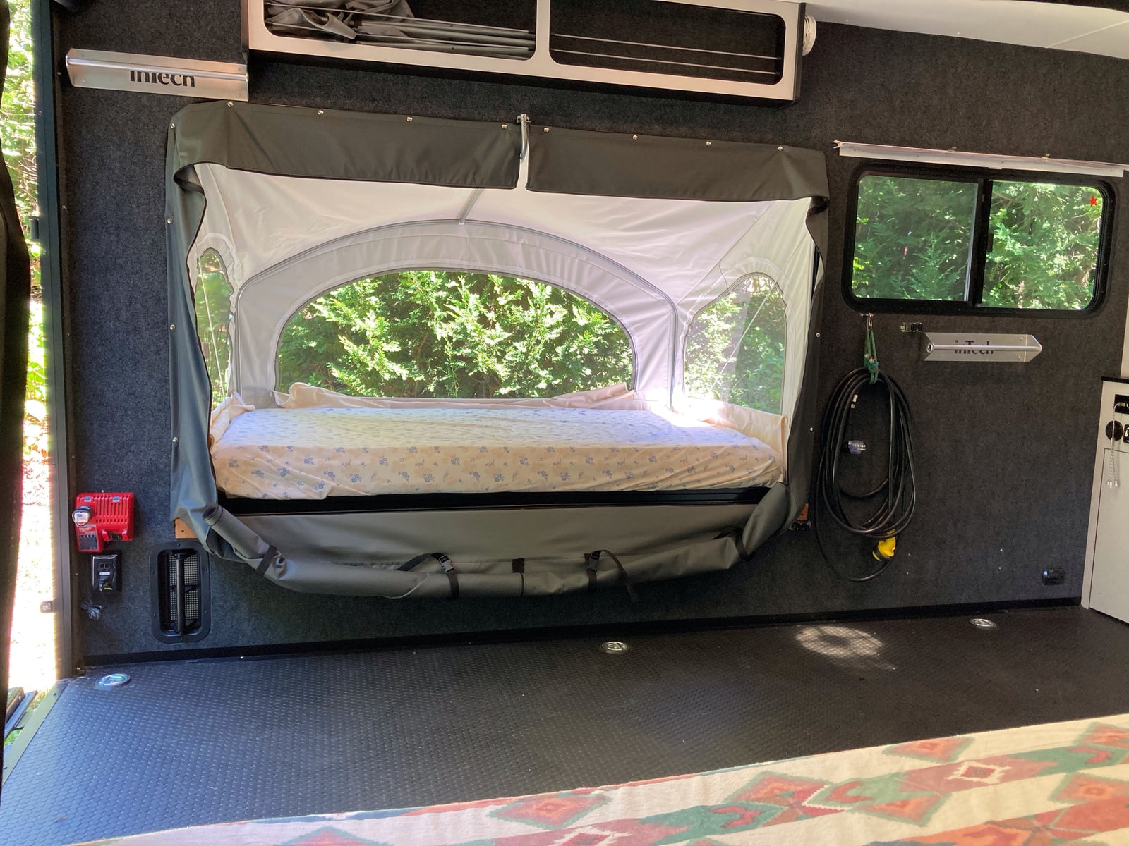 RV rental interior: a comfy pop-out tent bed with a mattress, windows showing lush trees, and a textured floor.