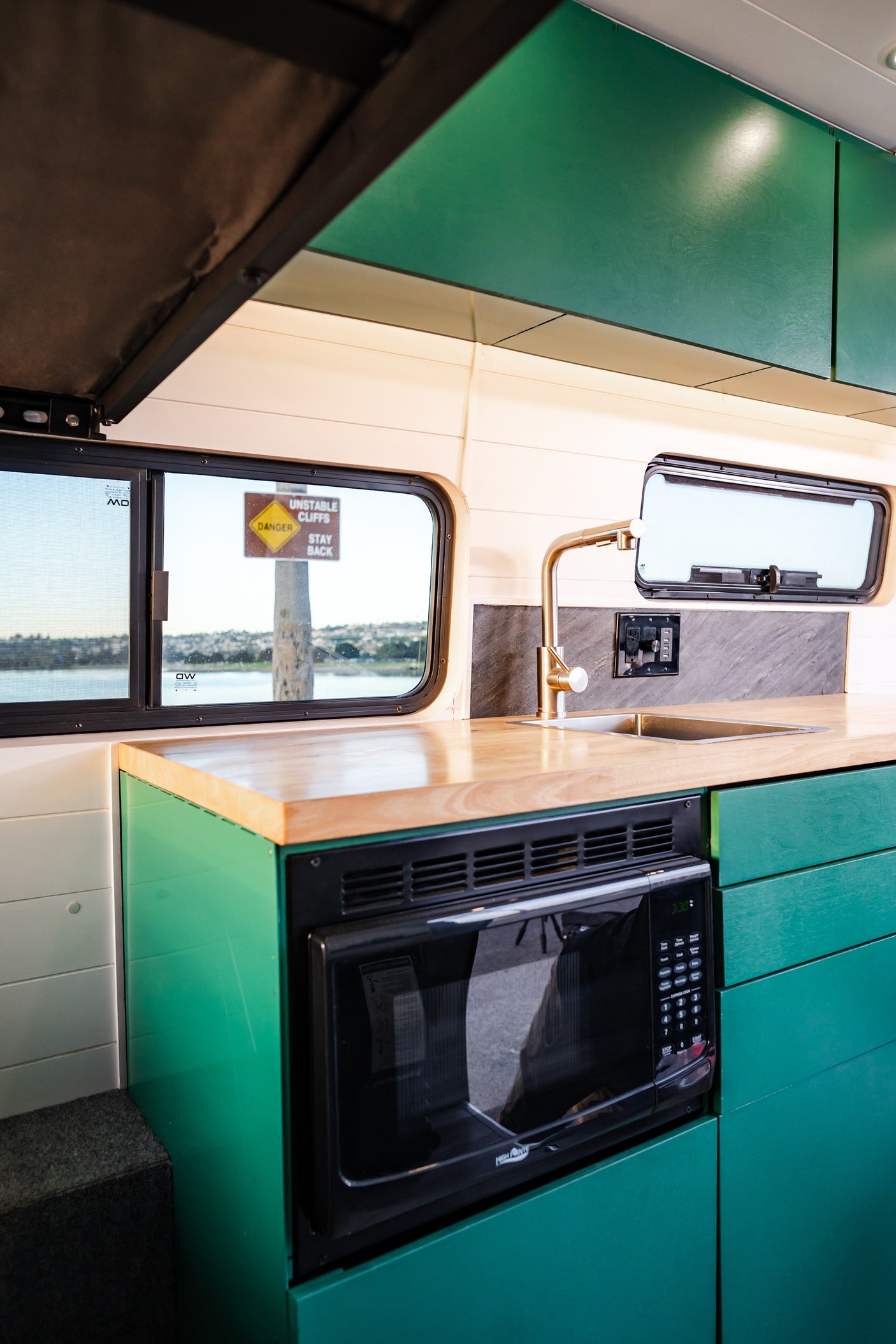 Compact RV rental kitchen with green cabinets, wood counter, sink, microwave. Window view: 'UNSTABLE CLIFFS' sign, water.