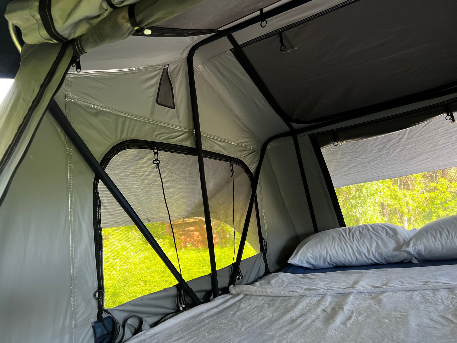 Cozy rv rental rooftop tent interior with white bedding, open windows, and a vibrant green nature view for outdoor adventures.