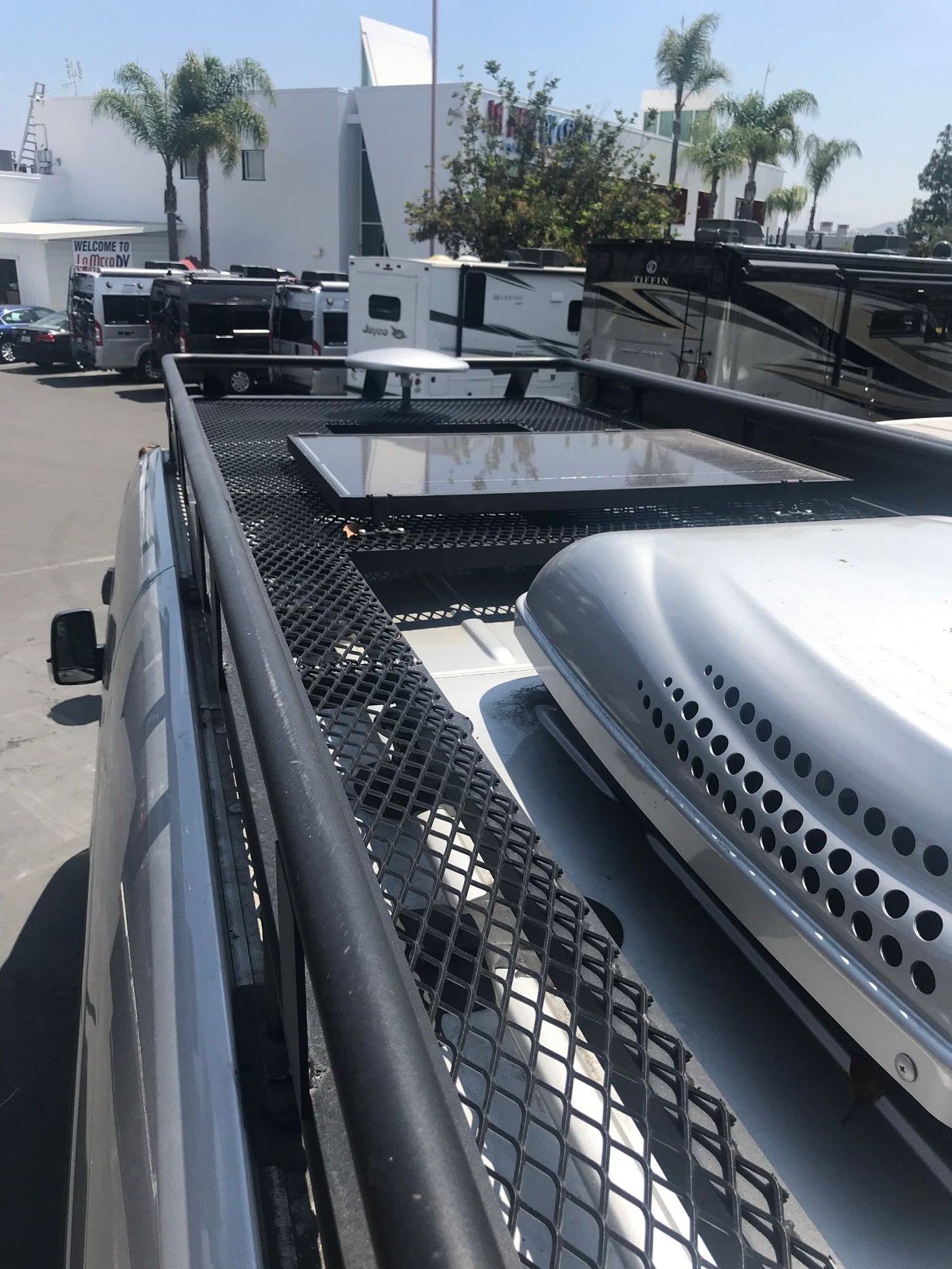 Rooftop view: solar panels, AC unit on an RV. Behind it, a sunny rv rental lot with diverse vans & motorhomes.