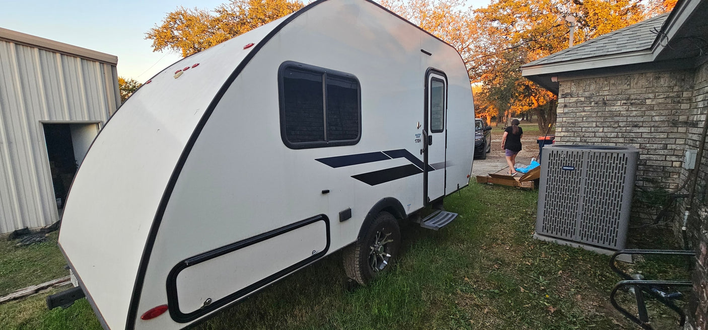 A white travel trailer RV rental with black accents is parked on a grassy yard between a metal shed and a brick home.|This white teardrop RV rental is parked on grass near a house