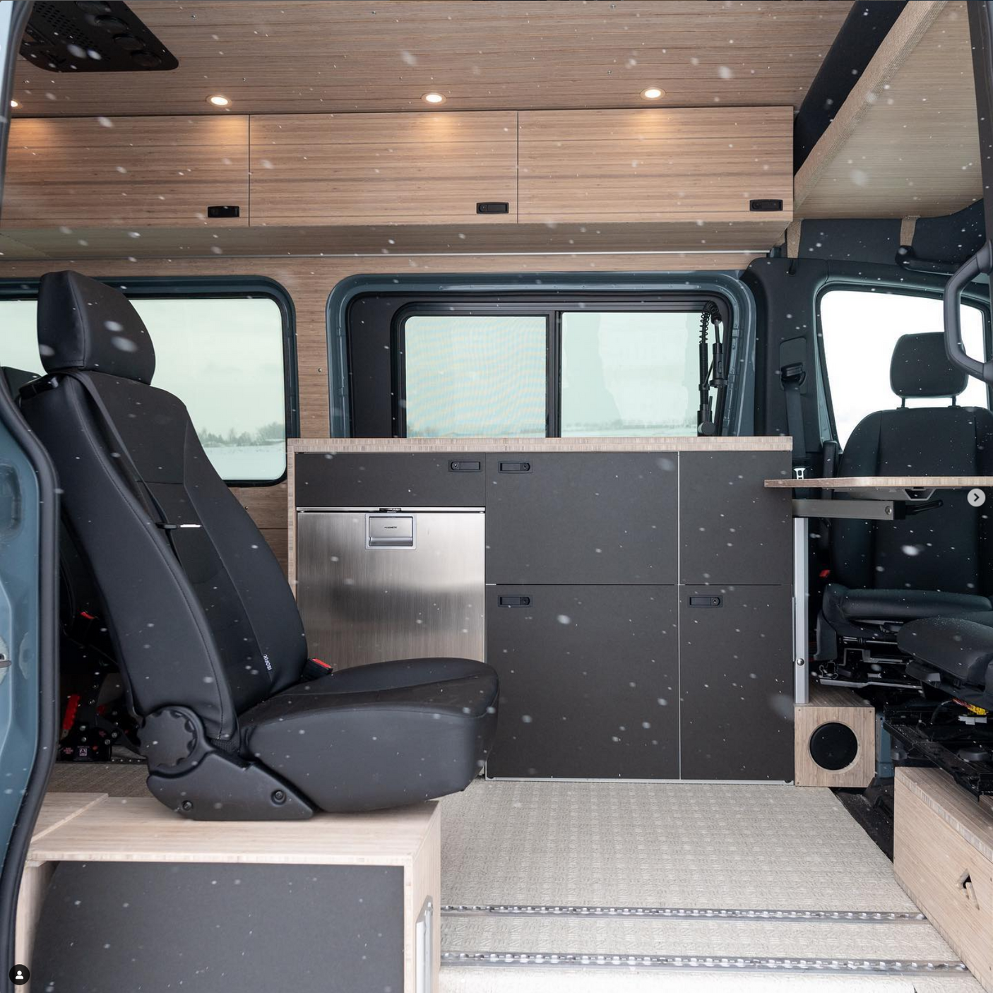 Modern RV rental interior with wood cabinetry, integrated Dometic fridge, comfy seating, and dark grey kitchen unit. Snowflakes dot the scene.