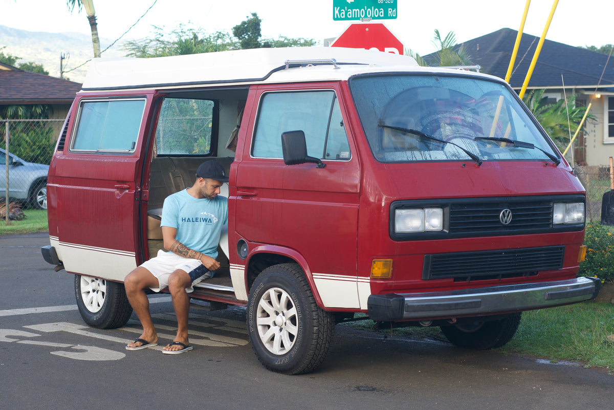 Man relaxes in an open red Volkswagen camper van, perfect for an RV rental. Parked on Ka'amōloa Rd.