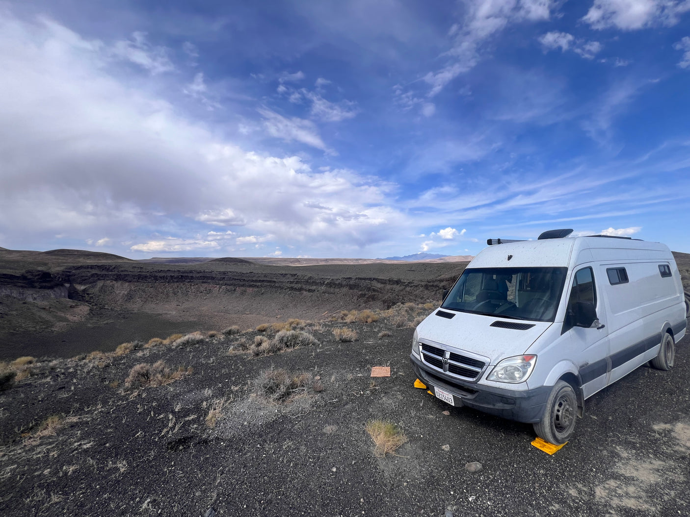 A white RV rental is parked on dark, rocky terrain, overlooking a massive volcanic crater with distant mountains under a cloudy sky.
