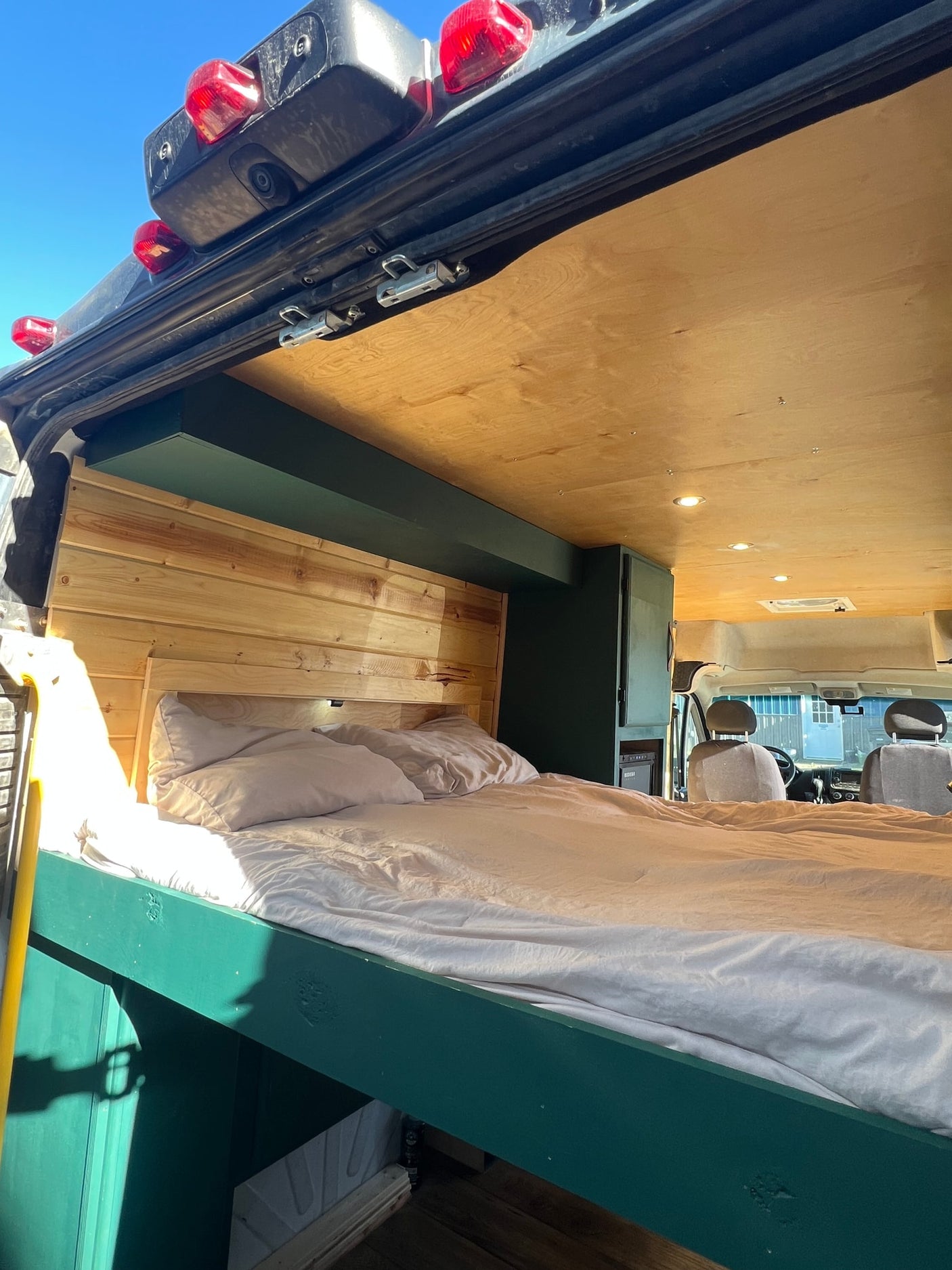 Bright RV rental interior: cozy bed with wood headboard, green frame, wood ceiling, and front cabin view.
