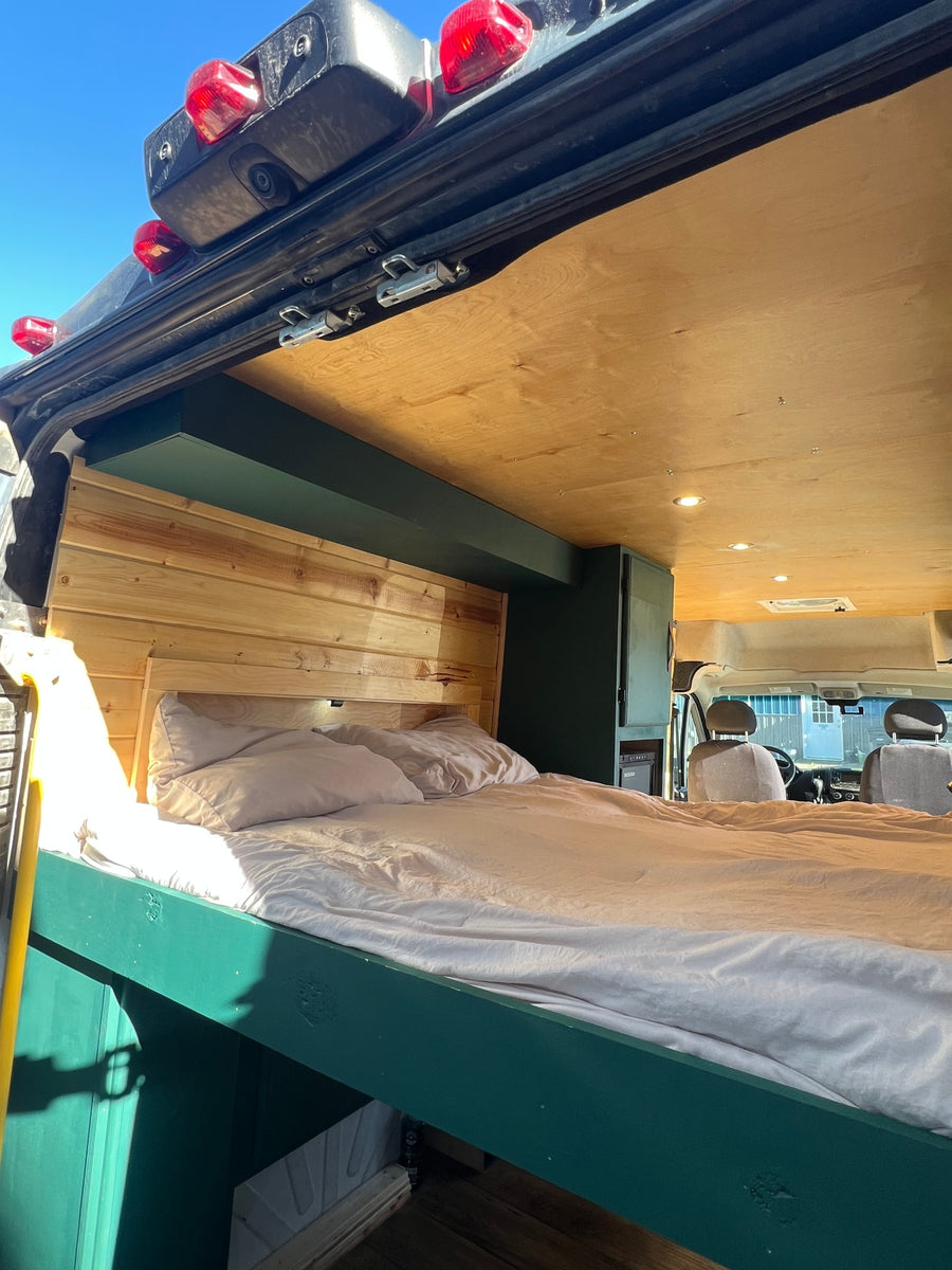 Bright RV rental interior: cozy bed with wood headboard, green frame, wood ceiling, and front cabin view.