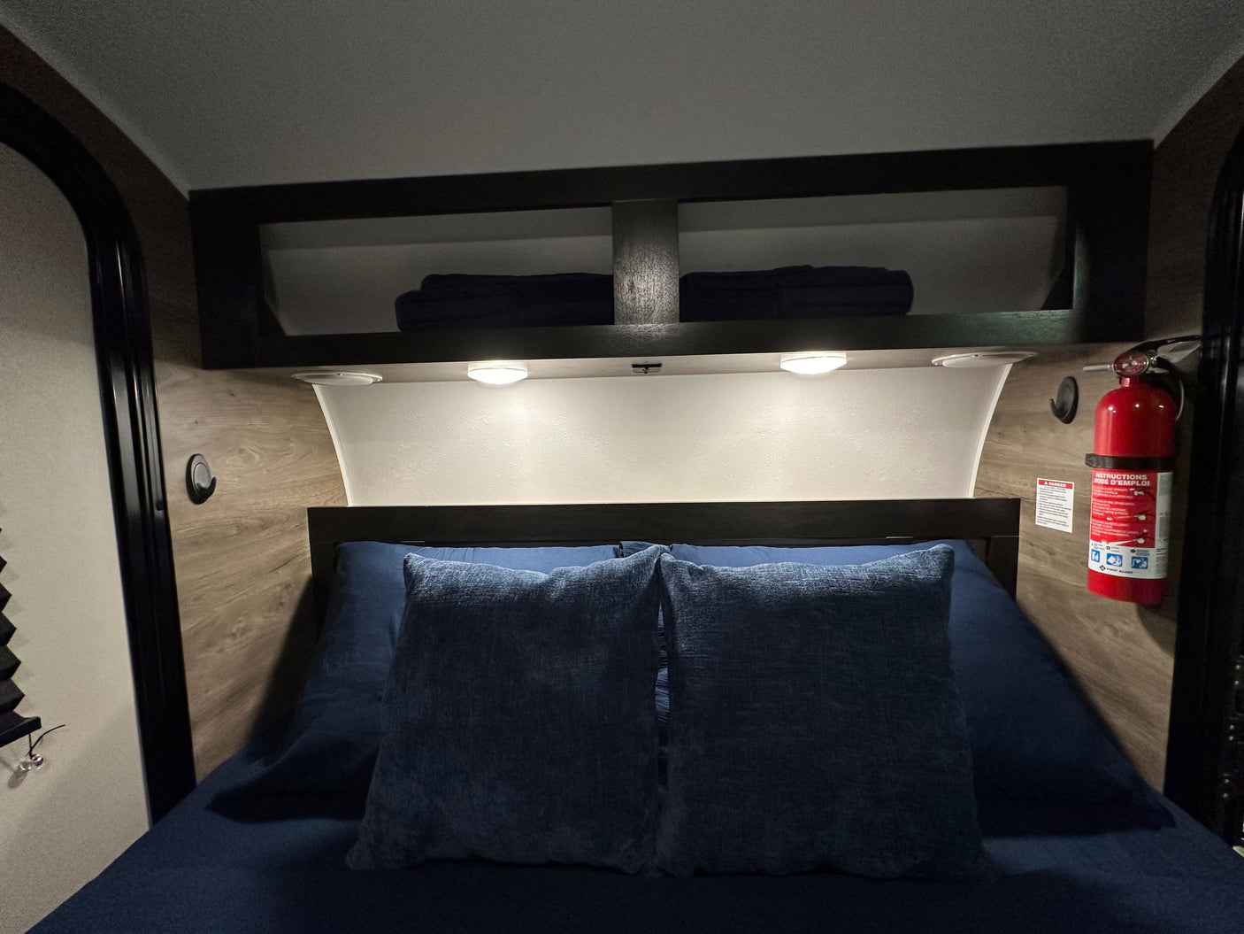 RV rental's cozy bedroom features a blue bed, overhead storage, and a visible fire extinguisher for safety.