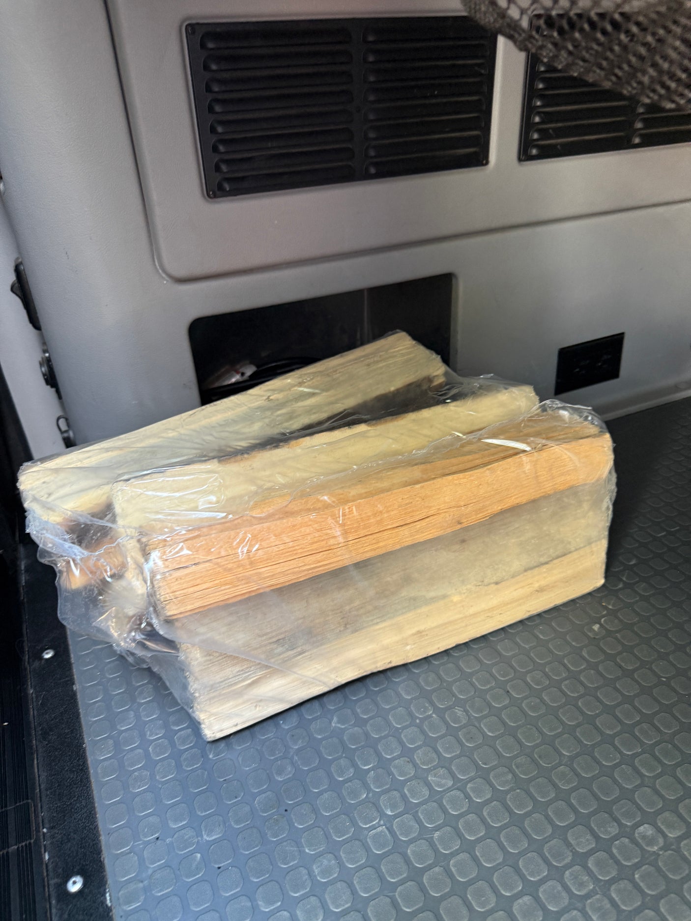 Bundled firewood, wrapped in plastic, rests on a textured floor mat in an RV rental interior, ready for a camping trip.