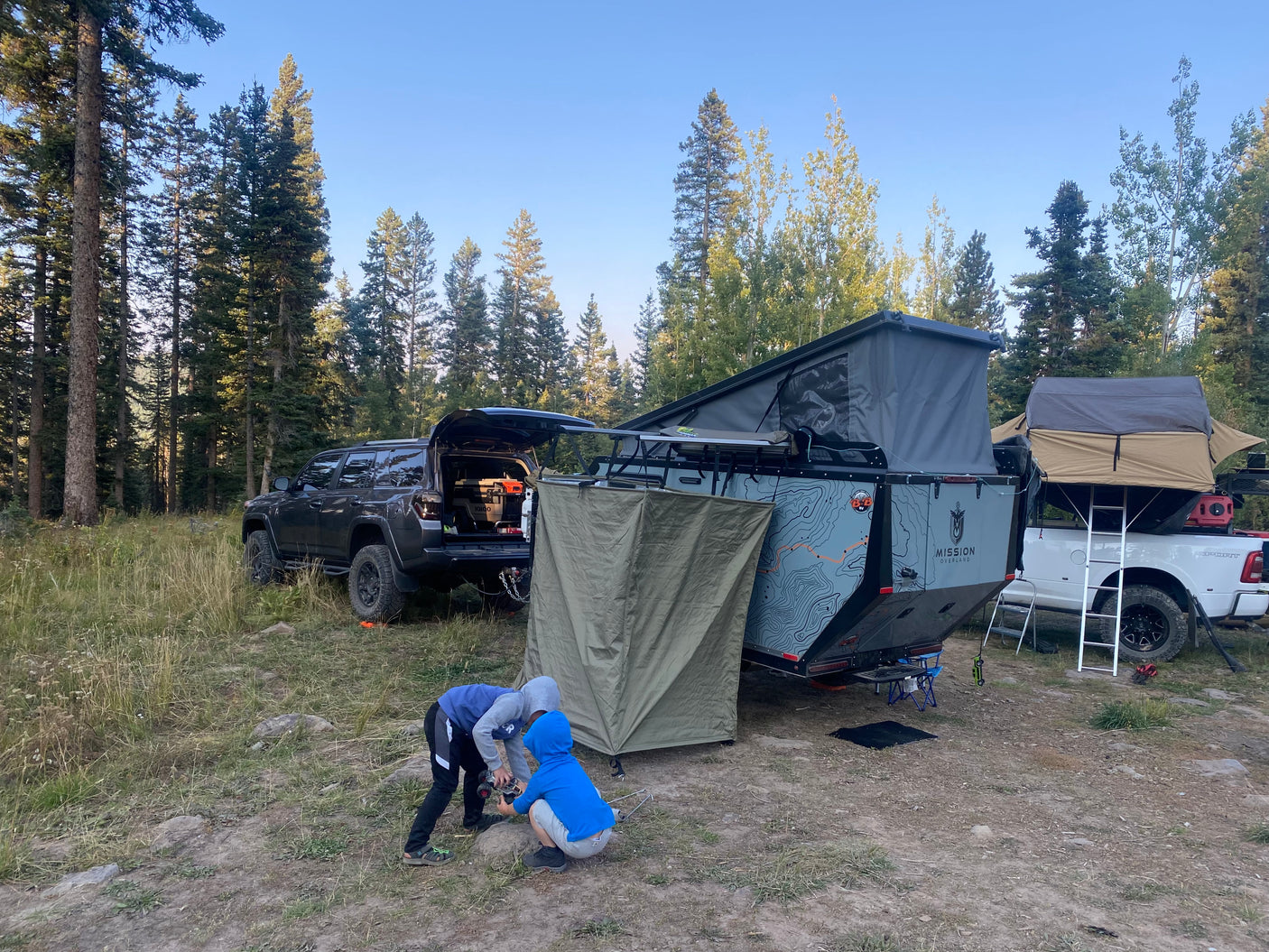 Campsite in a forest with an SUV, teardrop trailer, and truck with rooftop tent. Great for an RV rental adventure as kids play.