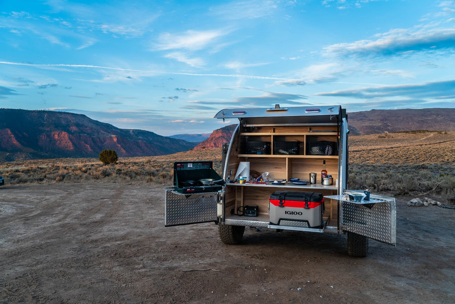 This rv rental teardrop trailer's open kitchen features a stove & cooler, ready for camping amidst scenic mountains. Perfect for adventure.