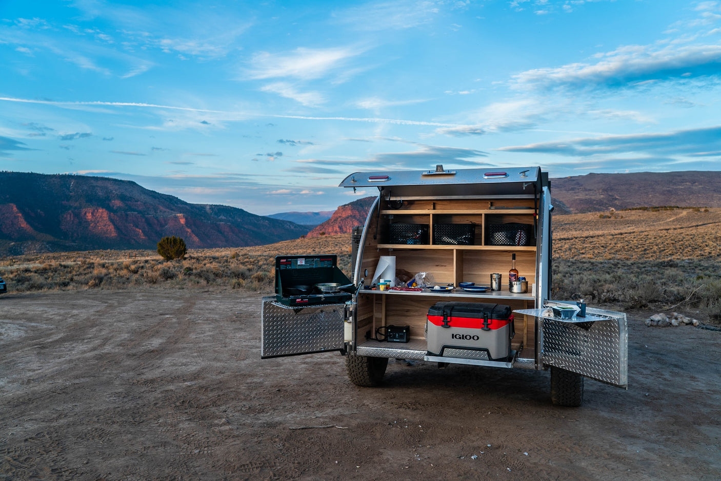 This rv rental teardrop trailer's open kitchen features a stove & cooler, ready for camping amidst scenic mountains. Perfect for adventure.