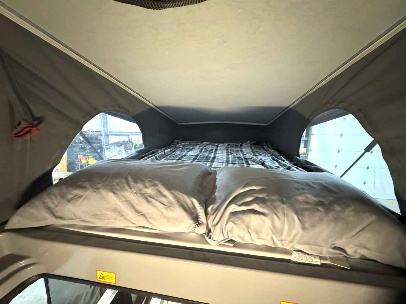 Interior of an RV rental pop-top tent: a made bed with pillows & blanket, windows, and a warning for adult supervision.