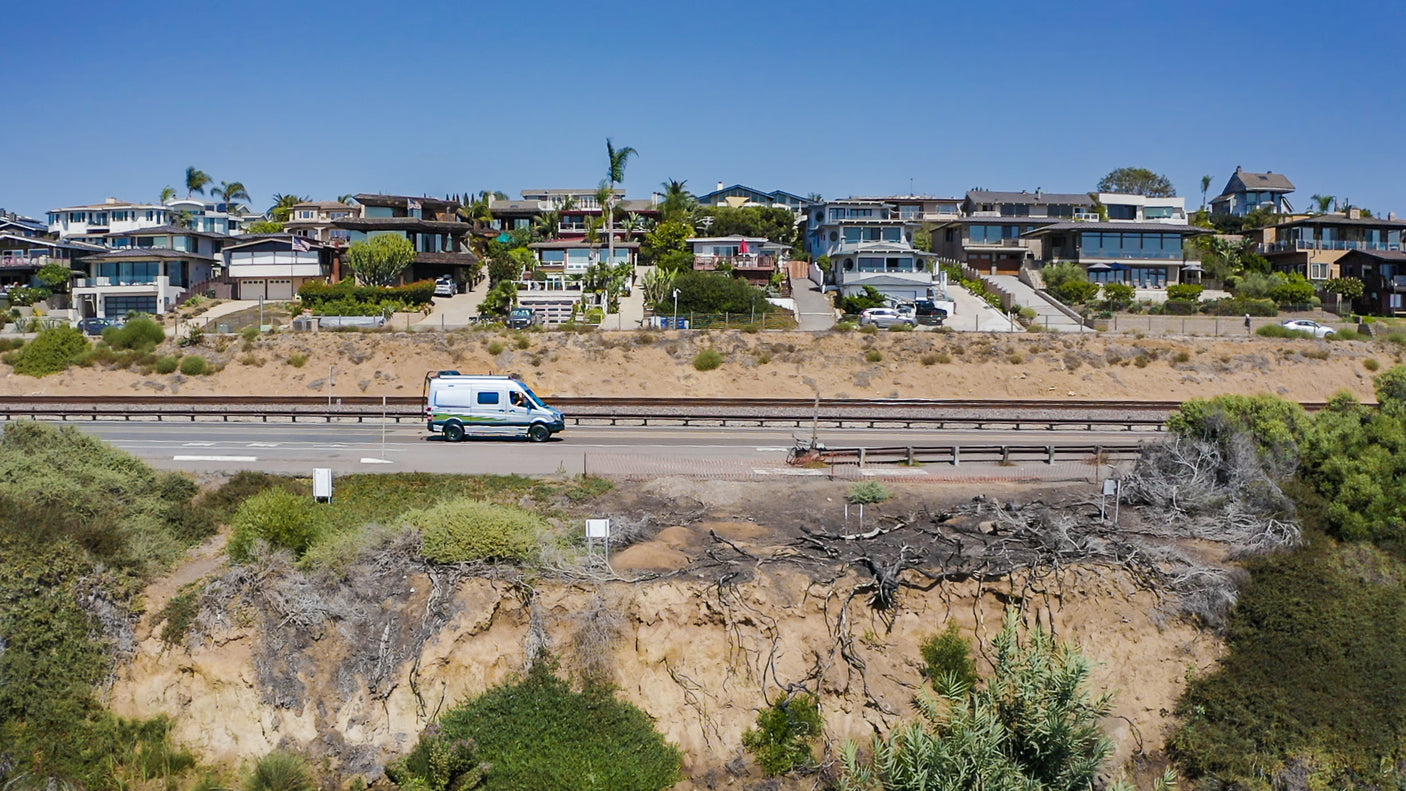White & green RV rental on coastal road, train tracks, hillside homes, and dry cliffs under a clear sky.