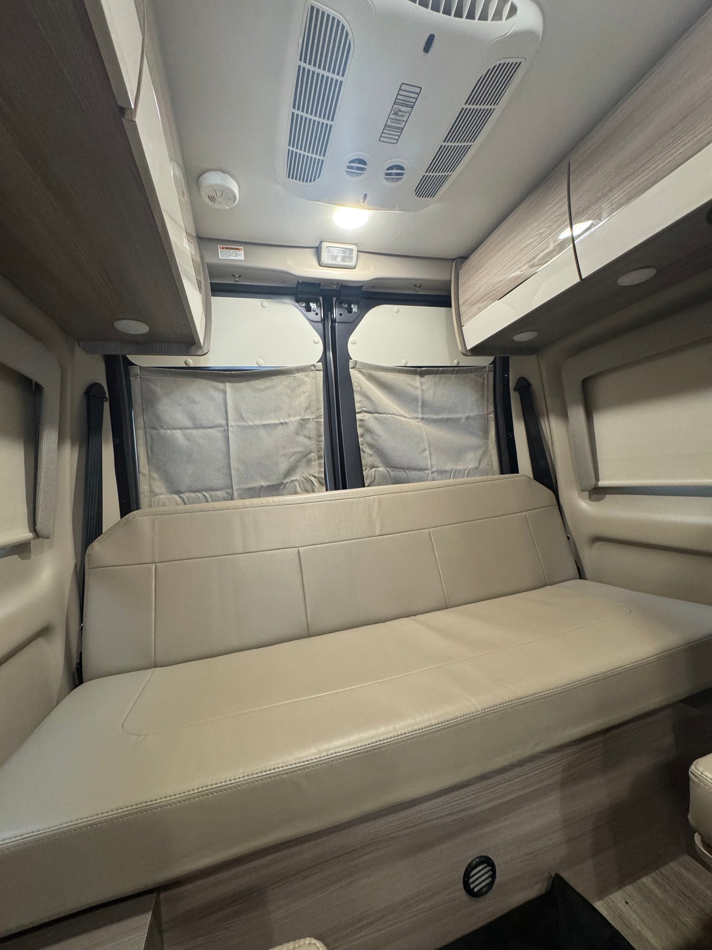 Spacious RV rental interior featuring a comfortable light-colored bench seat/bed, overhead cabinets, and ceiling AC unit.